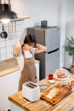 Pozitif, güzel bir Asyalı kadın yemek pişirirken, tost yaparken ve sabah kahvaltısını mutfakta hazırlarken saçını bağlıyor. Ev hayatı, ev hayatı, yemek ve insanlar.