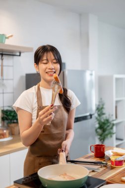 Önlüklü güzel, mutlu bir Asyalı kadın mutfakta kahvaltı hazırlıyor, tavada sosis pişiriyor. Ev yemeği, ev hayatı, insanlar, yemek.