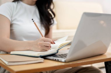 Konforlu giysiler içinde Asyalı bir üniversite öğrencisi, oturma odasında oturup mini bir masada ödev yapıyor, bir kitaba yazıyor, bilgisayarında çalışırken notlar alıyor.