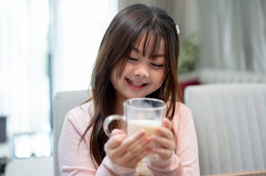 Genç, tapılası bir Asyalı çocuk kahvaltı sırasında yemek masasında bir bardak sütün tadını çıkarıyor..
