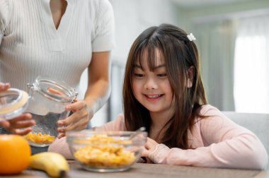 Genç, tapılası bir Asyalı kız yemek masasında oturuyor, sabah kahvaltılık gevreğini yemek için heyecanlanıyor. Şefkatli bir anne kızı için bir kavanozdan mısır gevreği dolduruyor..