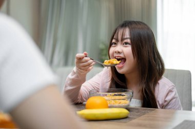 Genç, mutlu ve tatlı bir Asyalı kız annesiyle yemek masasında kahvaltı ederken, bir kase mısır gevreği yerken ve sabahları onunla kaliteli zaman geçirirken sohbet ediyor..