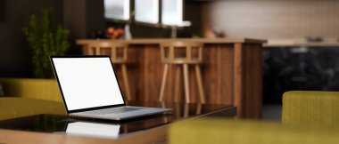 Çağdaş bir oturma odasındaki kahve masasının üzerinde beyaz ekran maketi olan bir dizüstü bilgisayarın yakın çekimi. Evin arka planında açık mutfak alanı var. 3d görüntüleme, 3d illüstrasyon