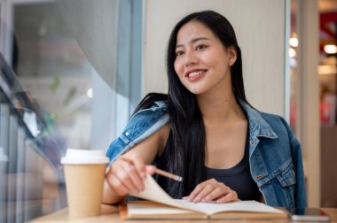Güzel, düşünceli Asyalı bir kadın kafenin penceresinde oturmuş günlüğüyle dışarıda bir şeyler düşünüyor ya da hayal kuruyor..