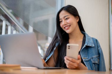 Uzaktan kumandalı bir kafede çalışan güzel bir Asyalı kadın dizüstü bilgisayarı üzerinde çalışırken akıllı telefonunu tutuyor. İnsanlar, yaşam tarzları, kablosuz teknoloji