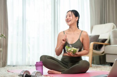 Yoga kıyafeti giymiş çekici, sıska bir Asyalı kadın yoga minderinde elinde sağlıklı bir salata kasesiyle oturmuş evde yoga yaptıktan sonra salata yiyor. ev hayatı, refah, sağlıklı hayat.