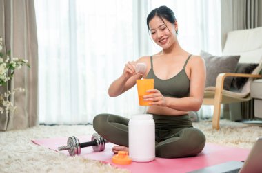 Yoga kıyafetleri içinde çekici, sağlıklı bir Asyalı kadın yoga minderinde oturmuş, idmandan sonra bir kepçe protein tozu ekleyerek protein içeceği hazırlıyor..