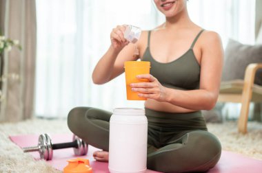 Yoga kıyafetleri içinde çekici, sağlıklı bir Asyalı kadının yakın plan fotoğrafı yoga minderinde oturup, çalışmasından sonra şişeye bir kepçe protein tozu ekleyerek protein içeceği hazırlıyor..