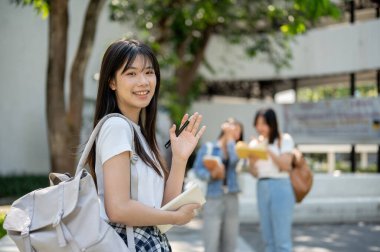 Hoş, arkadaş canlısı Asyalı bir üniversite öğrencisi. Günlük giysiler içinde ve sırt çantalı. Kolej parkında dikilirken kameraya gülümseyen ve elini sallayan iki kız..