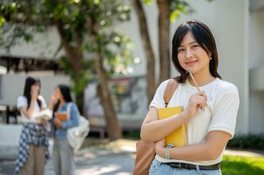Kolej parkında durmuş arka planda sohbet eden iki kızla kameraya gülümseyen, sıradan kıyafetler giymiş Asyalı tatlı bir üniversite öğrencisi. eğitim kavramı