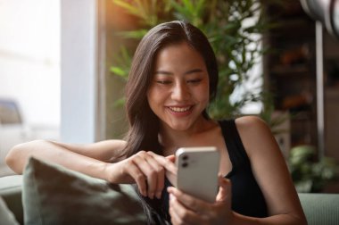 Güzel bir Asyalı kadının portresi. Kanepede dinleniyor. Akıllı telefonuyla sohbet etmekten zevk alırken gülümsüyor. İnsanlar, yaşam tarzları, kablosuz teknoloji