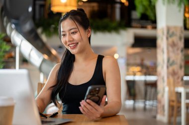 Güzel bir Asyalı kadın bir kahve dükkanında uzaktan çalışıyor, bilgisayarını tamir ederken akıllı telefonunu tutuyor. İnsanlar, yaşam tarzları, kablosuz teknoloji