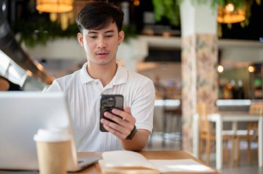 Yakışıklı bir Asyalı akıllı telefonuyla sohbet mesajları okumaya odaklanır. Bir kahve dükkanından uzaktan kumandayla, kapalı bir masada laptopuyla otururken..