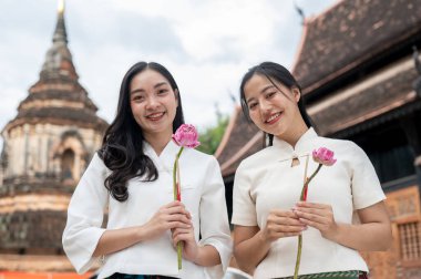 Geleneksel Kuzey Thai-Lanna elbiseli iki güzel Asyalı kadın bir tapınakta duruyorlar, ellerinde pembe nilüferler ve kameraya gülümsüyorlar. Tayland kültürü, Asya seyahati, Budist Tatilleri