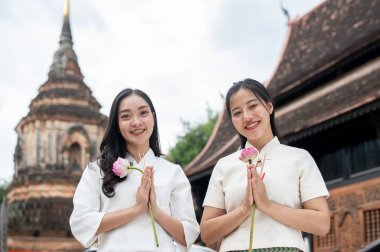 Geleneksel Kuzey Thai-Lanna elbiseli iki güzel Asyalı kadın bir tapınakta duruyorlar, ellerinde pembe nilüferler ve kameraya gülümsüyorlar. Tayland kültürü, Asya seyahati, Budist Tatilleri