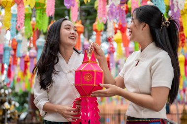 Geleneksel Kuzey Tayland-Lanna elbiseli iki çekici, gülümseyen Asyalı kadın bir tapınakta geleneksel bir fener festivalinin keyfini çıkararak bir arada tutuyorlar. Tayland kültürü, Budist Tatilleri