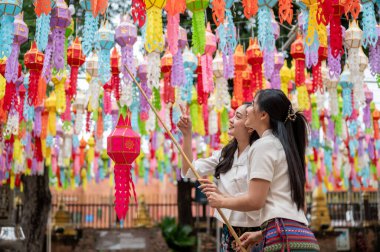 Geleneksel Kuzey Tayland-Lanna elbiseli iki büyüleyici Asyalı kadın tapınaktaki geleneksel bir fener festivalinde birlikte renkli bir fener asmaktan hoşlanıyorlar. Tayland kültürü, Budist festivali
