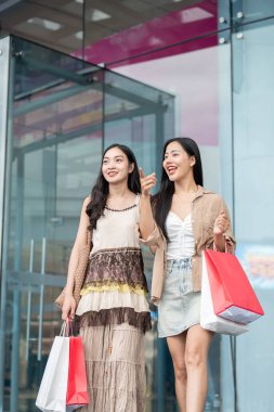 Moda kıyafetleri içinde iki mutlu, gülümseyen Asyalı bayan arkadaş alışveriş merkezinden alışveriş torbalarıyla çıkıp sokaktaki indirimli reklam panolarını göstererek kızların alışveriş günlerinin tadını çıkarıyorlar..