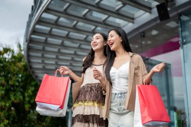 Moda kıyafetleri içinde iki çekici, neşeli Asyalı bayan arkadaş alışveriş merkezinin önünde durup alışveriş çantalarını taşıyorlar ve kameradan gülümsüyorlar..