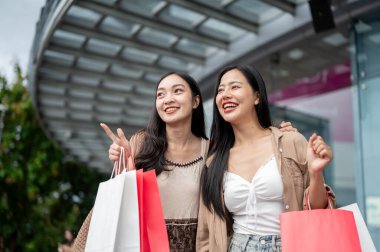 Moda kıyafetleri içinde iki mutlu, gülümseyen Asyalı bayan arkadaş alışveriş merkezinden alışveriş torbalarıyla çıkıp sokaktaki indirimli reklam panolarını göstererek kızların alışveriş günlerinin tadını çıkarıyorlar..