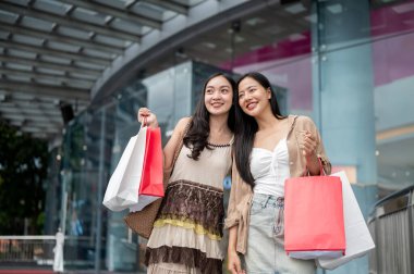 Moda kıyafetleri içinde iki çekici, neşeli Asyalı bayan arkadaş alışveriş merkezinin önünde durup alışveriş çantalarını taşıyorlar ve kameradan gülümsüyorlar..