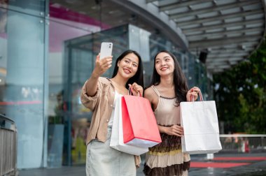 İki güzel, mutlu Asyalı bayan arkadaş alışveriş merkezindeki alışveriş çılgınlıklarından sonra akıllı telefonlarıyla selfie çekmeyi seviyorlar. Alışveriş çantalarını kameraya gösteriyorlar..