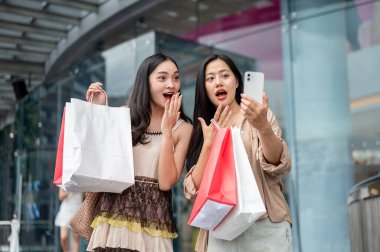 İki heyecanlı, neşeli Asyalı bayan arkadaş alışveriş merkezindeki alışveriş çılgınlıklarından sonra akıllı telefonlarıyla selfie çekiyorlar, alışveriş çantalarını ve sosyal medya paylaşımlarını gösteriyorlar..