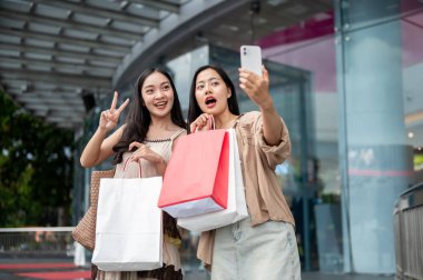 İki heyecanlı, neşeli Asyalı bayan arkadaş alışveriş merkezindeki alışveriş çılgınlıklarından sonra akıllı telefonlarıyla selfie çekiyorlar, alışveriş çantalarını ve sosyal medya paylaşımlarını gösteriyorlar..