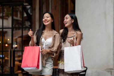 Moda kıyafetleri içinde iki güzel ve mutlu Asyalı kadın alışveriş torbalarıyla mağazadan çıkıp hafta sonu alışveriş çılgınlığının tadını çıkarıyorlar. şehir hayatı, seyahat, alışveriş, yaşam tarzları