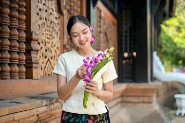 Geleneksel Kuzey Tayland elbisesi giymiş çekici bir Asyalı kadın elinde çiçeklerle tapınağın yanında duruyor. Budizm, maneviyat, Tayland kültürü.
