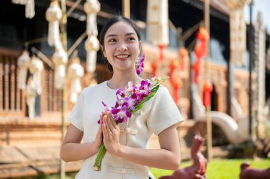 Geleneksel Kuzey Tayland elbisesi içinde güzel, çekici bir Asyalı kadın arka planda bir tapınakla dışarıda duruyor, kameradan uzağa gülümsüyor ve dua eden bir duruşla çiçekleri tutuyor..