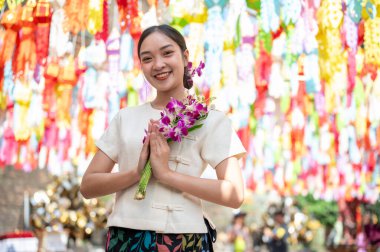 Geleneksel Kuzey Tayland elbisesi giymiş çekici bir Asyalı kadın tapınakta duruyor, kameraya gülümsüyor, elinde renkli fenerlerle bir buket orkide tutuyor. Loy Krathong festivali