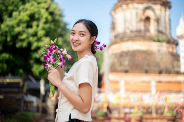Geleneksel Tayland Lanna elbisesi giymiş çekici bir Asyalı kadın elinde orkide çiçekleri arka planda güzel bir tapınak geçidiyle kameraya gülümsüyor. Tayland kültürü