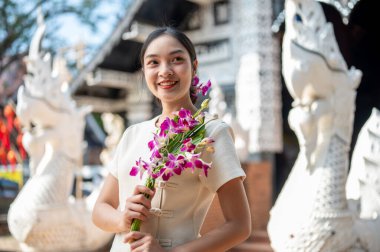 Geleneksel Tayland Lanna elbisesi giymiş çekici bir Asyalı kadın güzel bir antik tapınağın önünde duruyor, elinde bir orkide çiçeği buketi, kameradan uzağa bakıyor. Tayland Kültür Seyahati