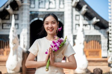Geleneksel Tayland Lanna elbisesi giymiş çekici bir Asyalı kadın dua eden bir duruşla elinde çiçeklerle güzel bir tapınağın önünde kameraya bakıyor. Tayland Kültür Seyahati