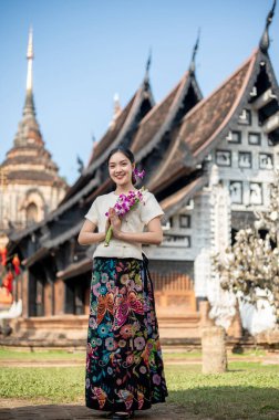 Geleneksel Tayland Lanna elbisesi giymiş çekici, gülümseyen Asyalı bir kadın arka planda güzel bir tapınak ve pagoda ile dışarıda duruyor. Elinde dua eden bir çiçek buketi var..