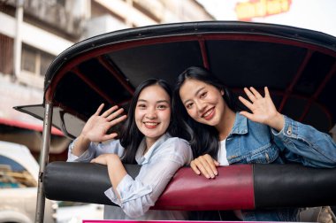 İki neşeli, çekici Asyalı genç kız Tayland 'da bir tuk-tuk' ta kameraya el sallayıp gülümsüyorlar. Turist, seyahat, ulaşım, Tayland