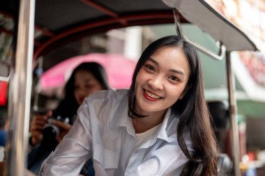 Güzel, gülümseyen Asyalı genç bir bayan turist arkadaşıyla Tayland 'da bir tuk-tuk' ta kameraya bakarak eğleniyor. Turizm, seyahat, ulaşım