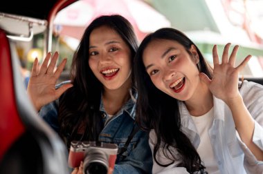İki neşeli, çekici Asyalı genç kız Tayland 'da bir tuk-tuk' ta kameraya el sallayıp gülümsüyorlar. Turist, seyahat, ulaşım, Tayland