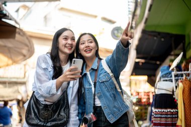İki heyecanlı, neşeli Asyalı bayan arkadaş yerel bir pazarı birlikte keşfetmekten zevk alıyorlar, bir yeri araştırıyorlar, akıllı telefondan haritaya bakıyorlar, bir yemek standını işaret ediyorlar, Güneydoğu Asya 'da eğleniyorlar.