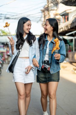 İki çekici, mutlu Asyalı bayan arkadaş, güneşli bir günde Tayland 'da sokak yemeği yiyerek ve kahkahalarını paylaşarak şehir gezintisi yapıyorlar. Tayland tatili, yaz gezisi, turizm
