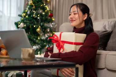 Güzel, neşeli Asyalı bir kadın oturma odasında oturmuş dizüstü bilgisayarından erkek arkadaşına Noel hediyesi kutusunu gösteriyor. Noel tatili kavramı