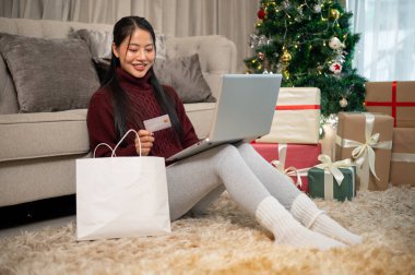 Mutlu bir Asyalı kadın Noel tatili boyunca evde online alışveriş yapmaktan hoşlanır. Oturma odasında dizüstü bilgisayarını kullanır ve sıcak, şenlikli bir Noel atmosferiyle çevrilidir..