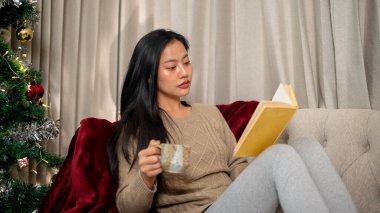 Sıcak, rahat giysiler içinde rahat bir Asyalı kadın kitap okuyor ve Noel gecesi huzurlu bir atmosferde, oturma odasındaki kanepede sıcak bir şeyler içiyor..