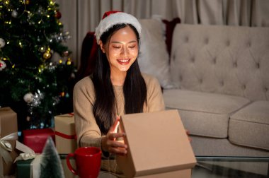 Güzel, mutlu, rahat elbiseli Asyalı bir kadın Noel hediyesini sıcak bir Noel atmosferiyle çevrili oturma odasında açıyor..