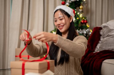 Çekici, gülümseyen Asyalı bir kadın Noel hediyelerini oturma odasında hazırlıyor, hediye kutusuna kırmızı bir kurdele bağlıyor. tatil, Noel gecesi, kutlama