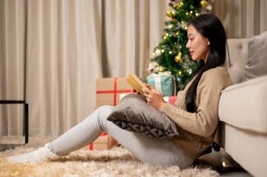 Güzel, mutlu, sıcacık giysiler içinde Asyalı bir kadının yan görüntüsü Noel gecesi oturma odasında kitap okumaktan ve Noel tatilini evde yalnız geçirmekten zevk alıyor..