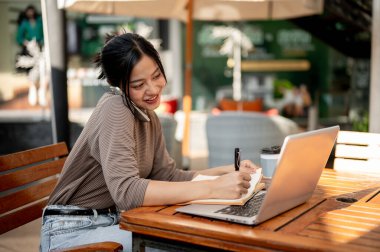 Güzel, çekici bir Asyalı kadın telefonda konuşurken bir kitaba not alıyor ve şehirdeki bir kafede bilgisayarıyla dışarıda bir masada oturuyor..