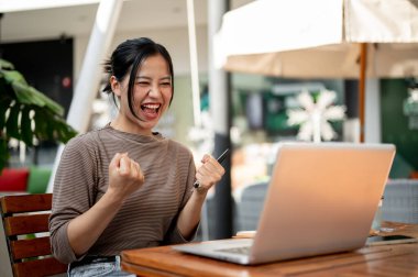Neşeli Asyalı bir kadın dışarıda dizüstü bilgisayarının önünde oturur, mutlu bir ifadeyle ekrana bakar, zaferle yumruğunu kaldırır, başarısını heyecanlı bir sevinç çığlığıyla kutlar..