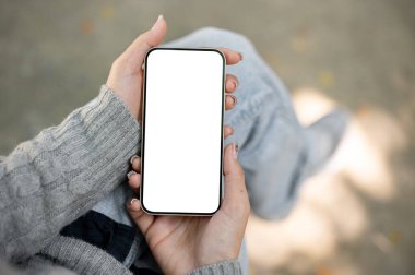 Bir kadının elinde beyaz ekran modeli olan bir akıllı telefonun yakın plan görüntüsü, bulanık bir arka plan üzerine kurulmuş. Kadın bir bankta oturuyor, akıllı telefonunu kullanıyor.